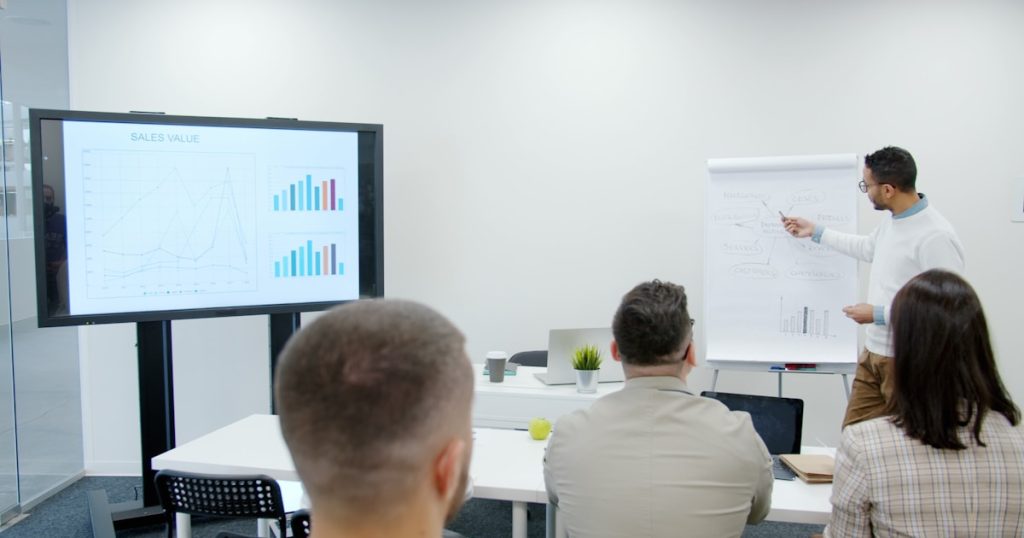 Man presents charts to seated audience in a modern office.